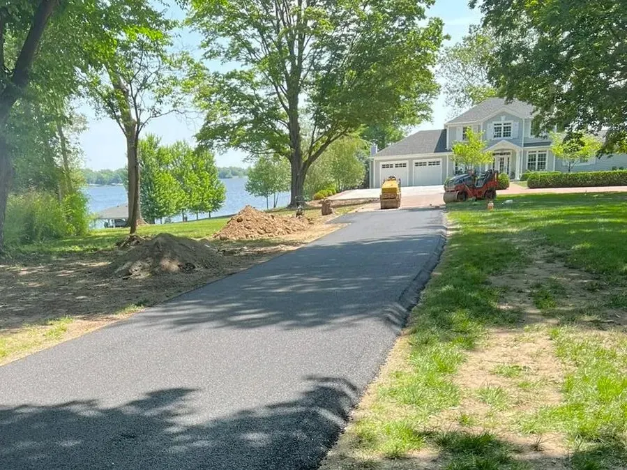 Newly paved asphalt driveway curving toward a lakeside home with rollers compacting the surface.